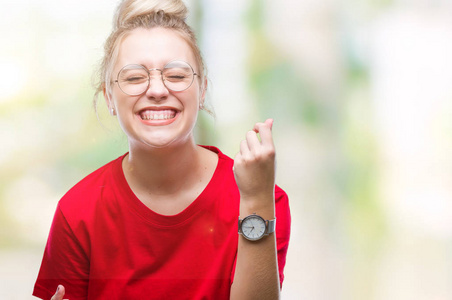 年轻的金发女人戴着眼镜，在孤立的背景下，非常高兴和兴奋地做着胜利者的手势，手臂抬起，微笑着，尖叫着，争取成功。 庆祝概念。