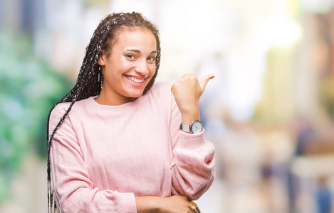 年轻的辫子非裔美国女孩穿着毛衣，在孤立的背景上微笑，快乐的脸，看着，用拇指指向一边。