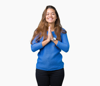 年轻漂亮的黑发女人穿着蓝色毛衣，在孤立的背景上祈祷，双手一起祈求宽恕，微笑自信。