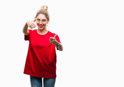 年轻漂亮的金发女人穿着红色的T恤和眼镜，在孤立的背景上，批准用手竖起大拇指做积极的手势，微笑着，为成功而高兴。 看着相机赢家的手