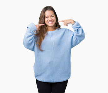 年轻漂亮的黑发女人穿着蓝色的冬季毛衣，在孤立的背景上微笑，自信地展示和指着手指，牙齿和嘴。 健康概念。