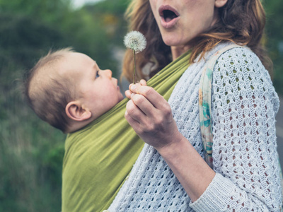 一位年轻的母亲带着一个婴儿在吊带里吹着蒲公英的种子