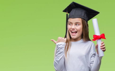 年轻漂亮的女孩戴着研究生帽，抱着学位，在孤立的背景下指着，用拇指向露出快乐的脸微笑