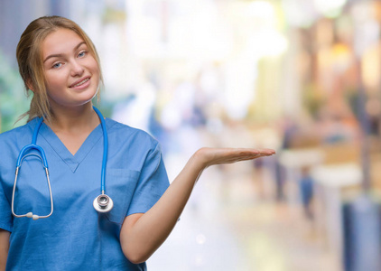 年轻的白种人医生女士穿着外科医生制服，在孤立的背景上微笑，欢快的呈现，用手掌指着相机。
