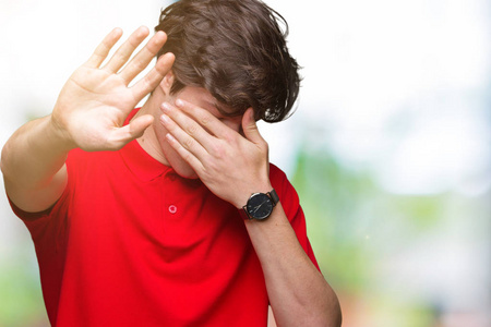 年轻的英俊男子穿着红色T恤，在孤立的背景上，用手遮住眼睛，用悲伤和恐惧的表情做停止手势。 尴尬和消极的概念。