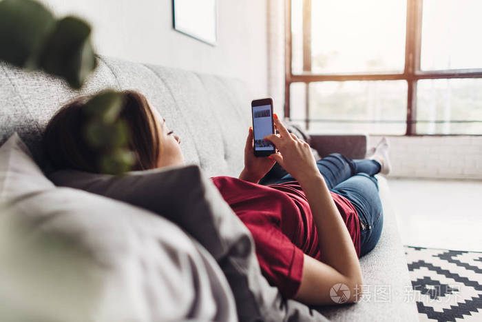 年轻的妇女使用智能手机躺在沙发上在家