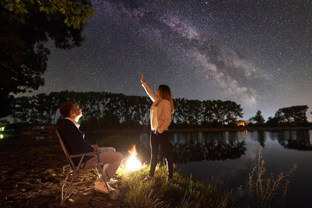 男人坐在椅子上，女人指着夜空，满是星星和银河