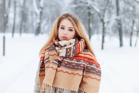 时尚迷人的年轻女子金发，穿着一条老式的羊毛围巾，在雪地公园的背景上穿着一件灰色外套，图案很漂亮。 户外时尚漂亮的女孩。