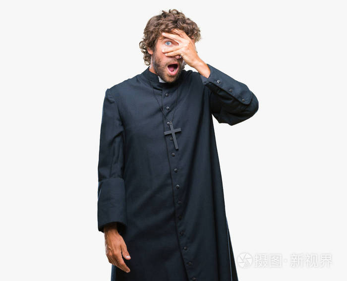 英俊的西班牙裔天主教牧师男子在孤立的背景上偷看震惊,覆盖着脸和