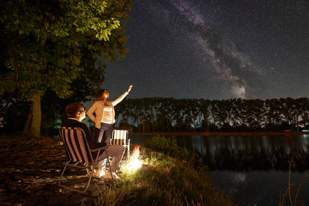男人坐在椅子上，女人指着夜空，满是星星和银河