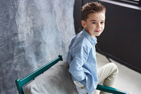 时髦的男孩穿着蓝色衬衫，坐在阁楼工作室的椅子上，灰色的背景。