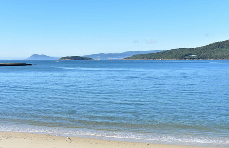 海滩在海湾与岛屿小海岸村庄和森林。 明亮的沙子和蓝色的大海，泡沫，晴天，晴空。 加利西亚西班牙。