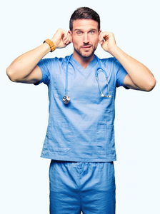 英俊的医生，穿着医疗制服，在孤立的背景上，用手指遮住耳朵，因为嘈杂的音乐，他的表情很生气。 聋的概念。