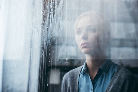 在家中悲伤的成年女人用雨滴透过窗户看着