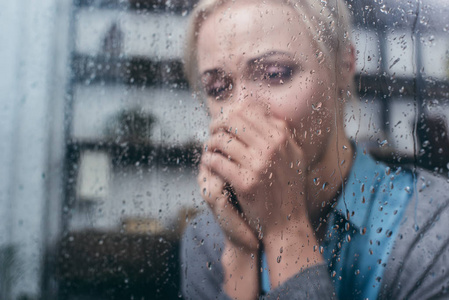 沮丧的成年女人，双手交叉在家里，透过窗户和雨滴