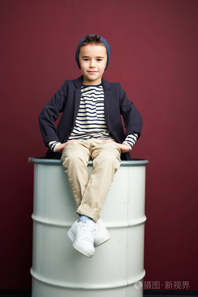 穿着时尚休闲服的男孩坐在白色桶上. 棕色背景工作室