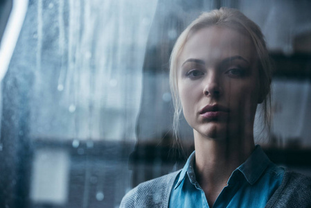 在家中悲伤的成年女人用雨滴透过窗户看镜头