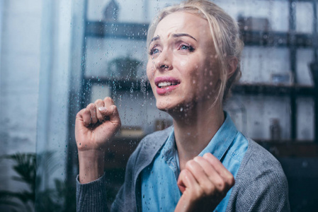 悲伤的成年女人，攥着拳头在家里哭，透过窗户，雨点打在她的身上