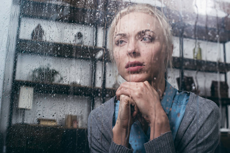 悲伤的成年女人，双手合拢，透过窗户看着家，雨滴