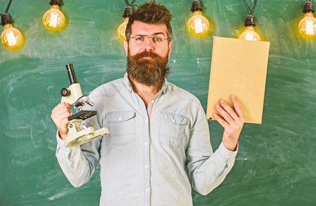 戴眼镜的老师手持书本和显微镜。有胡子和胡子的男人在教室里严脸。科学家拿着书和显微镜, 黑板上的背景, 复制空间。科学理念