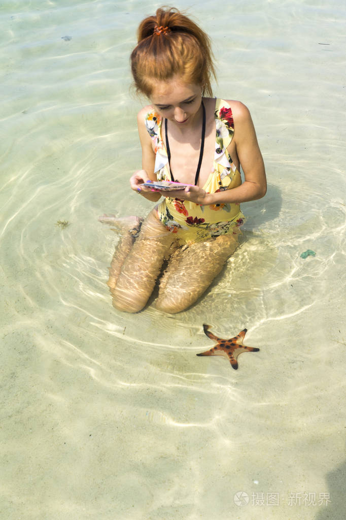 穿着比基尼的红发女孩,和星星鱼坐在一起,抱着椰子在普吉岛的海滩上