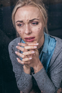 沮丧的成年女人，双手合拢，在家中哭泣，透过窗户，雨滴