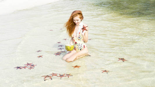 穿着比基尼的红发女孩，和星星鱼坐在一起，抱着椰子在普吉岛的海滩上，在一个晶莹剔透的海上旅行概念