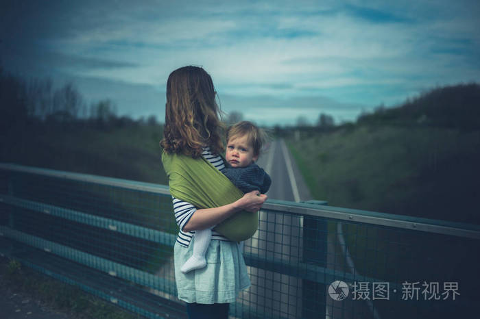一位年轻的母亲抱着蹒跚学步的孩子站在路上的桥上