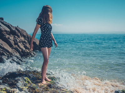 一位年轻女子站在海滩的岩石上，海浪拍打着。