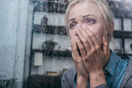 悲伤的成年女人哭着，用手捂着脸，透过窗户和雨滴