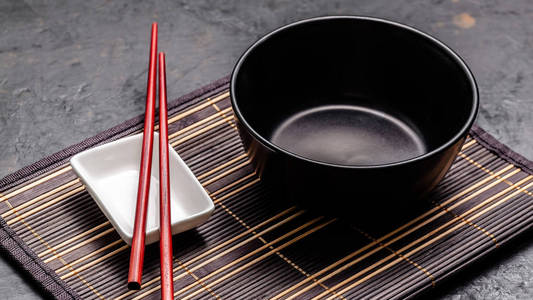 空的日本菜。 一个黑色的陶瓷碗，用于中国面条或泰国汤，放在一姆库克地毯上。 酱油白酱锅和红色中国棍子在黑色背景上。 顶部视图复制