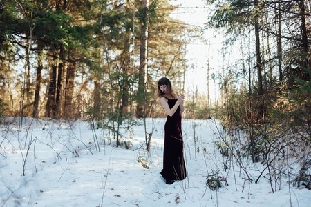 一个穿着深色长裙的美丽女孩的肖像，她独自站在冬天森林里的树木中。