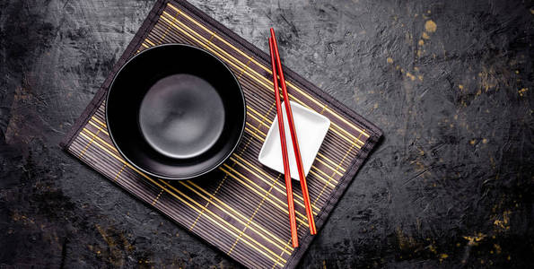 空的日本菜。 一个黑色的陶瓷碗，用于中国面条或泰国汤，放在一姆库克地毯上。 酱油白酱锅和红色中国棍子在黑色背景上。 顶部视图复制