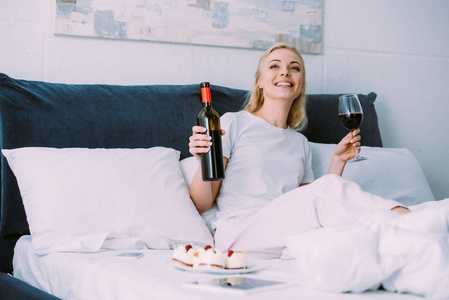 微笑的女人独自一人在床上庆祝生日时拿着瓶子和一杯红酒