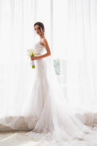 美丽幸福的新娘站在窗户附近，手里拿着一束花