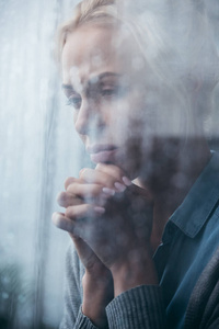 悲伤的成年女人，双手交叉在家，透过窗户，雨滴