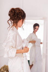 美丽的年轻新娘站在室内镜子前，手里拿着她的婚纱