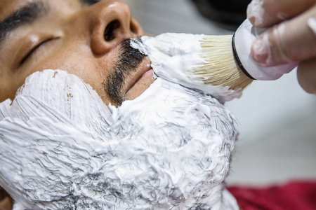 理发师用刷子在年轻的黑人胡须上涂抹白色剃须膏。理发师在理发店沙龙的顾客脸上涂抹泡沫。男性美容护理理念