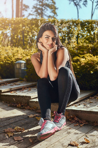 一个迷人的健身女孩, 皮肤晒黑, 穿着运动服, 坐在公园的木台阶上, 面带微笑, 看着相机