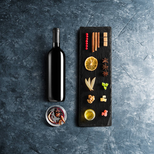 覆盖葡萄酒配方成分和厨房配件瓶红葡萄酒，肉桂，八角星，橙色红糖和香料在蓝色的石头背景。