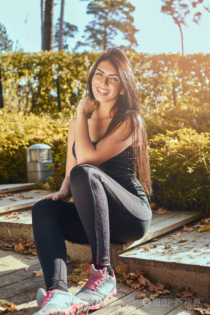 一个迷人的健身女孩, 皮肤晒黑, 穿着运动服, 坐在公园的木台阶上, 面