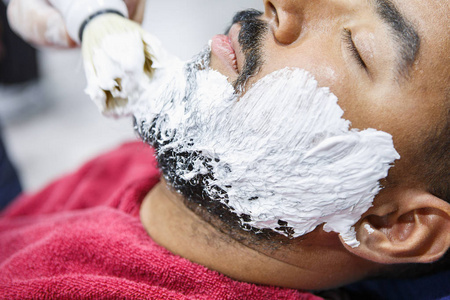 理发师用专业的拭子刷在年轻的黑人脸上涂上白色剃须膏，以便准确地刮胡子。男性美容治疗理念。