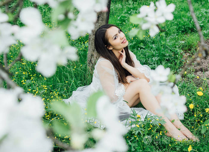 花苹果园里穿着白色裙子的微笑少女坐在草地上的画像