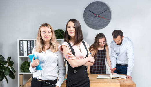 课题业务，团队合作及办公室内的谈判..一组4个白人在房间里拿文件工作。两位年轻的女老板在公司员工的背景下摆姿势。