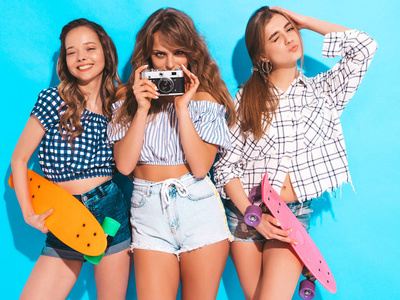 三个，美丽，时尚，微笑的女孩与五颜六色的硬币滑板。女人在夏季时髦的格子衬衫衣服摆在蓝色的墙壁附近。 模特们在复古相机拍照