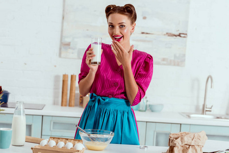漂亮的微笑别针女孩拿着一杯牛奶站在厨房桌子旁，有不同的产品