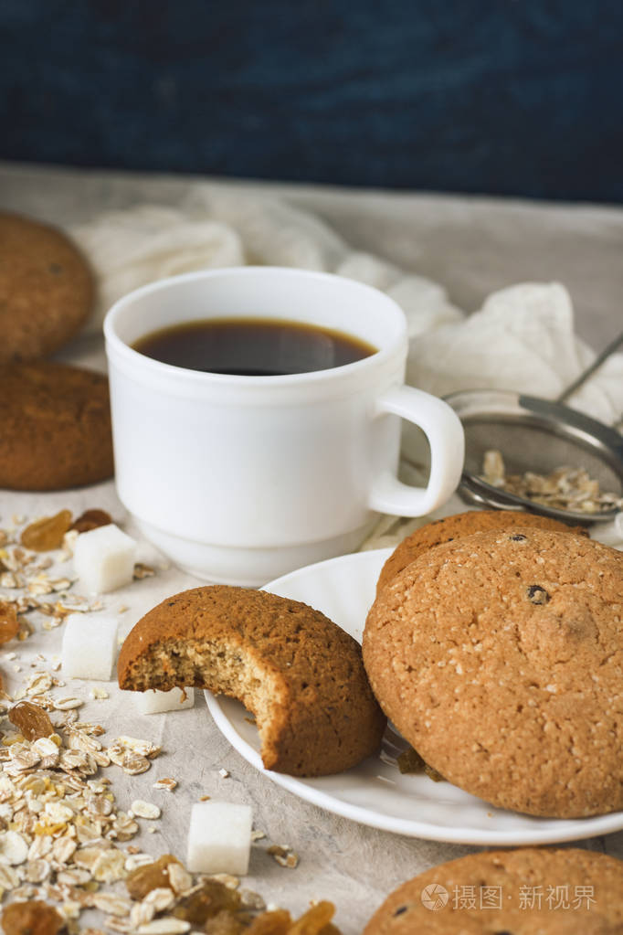 白色杯子中的黑咖啡杯燕麦饼干燕麦片葡萄干在浅色背景下早餐场景