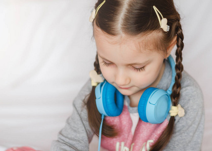 带着蓝色耳机的小女孩手里拿着一部智能手机，坐在床上。 穿着粉红色睡衣的小女孩。 选择性聚焦。 听音乐。