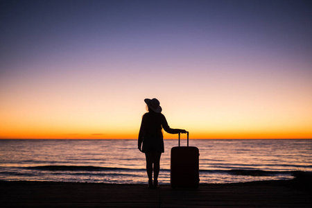 放松妇女与手提箱在海滩上日落剪影。假日旅行的概念。在海洋风景背景与手提箱的年轻夫人