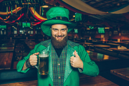 在酒吧里穿绿色西装的快乐而积极的年轻人。他拿着一杯啤酒, 竖起大拇指。年轻人很满意